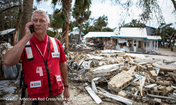 A Red Cross volunteer speaks on a phone in front of debris and destruction in the background. 