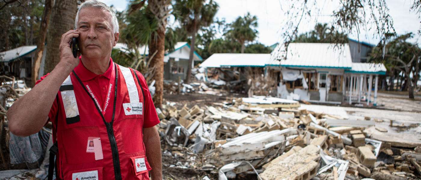 A Red Cross volunteer speaks on a phone in front of debris and destruction in the background. 