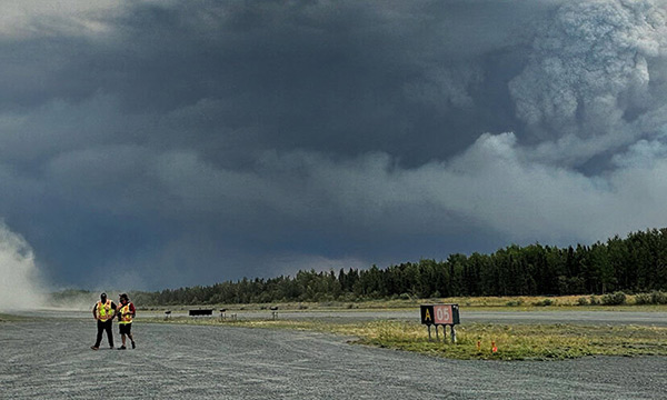 Two people in high-vis vests walk across an airfield as smoke rises from a forest behind them.