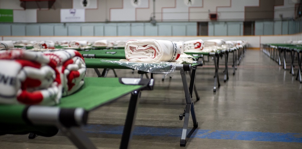 Rows of green cots with folded Red Cross blankets on them sit on the floor of a hockey arena. 