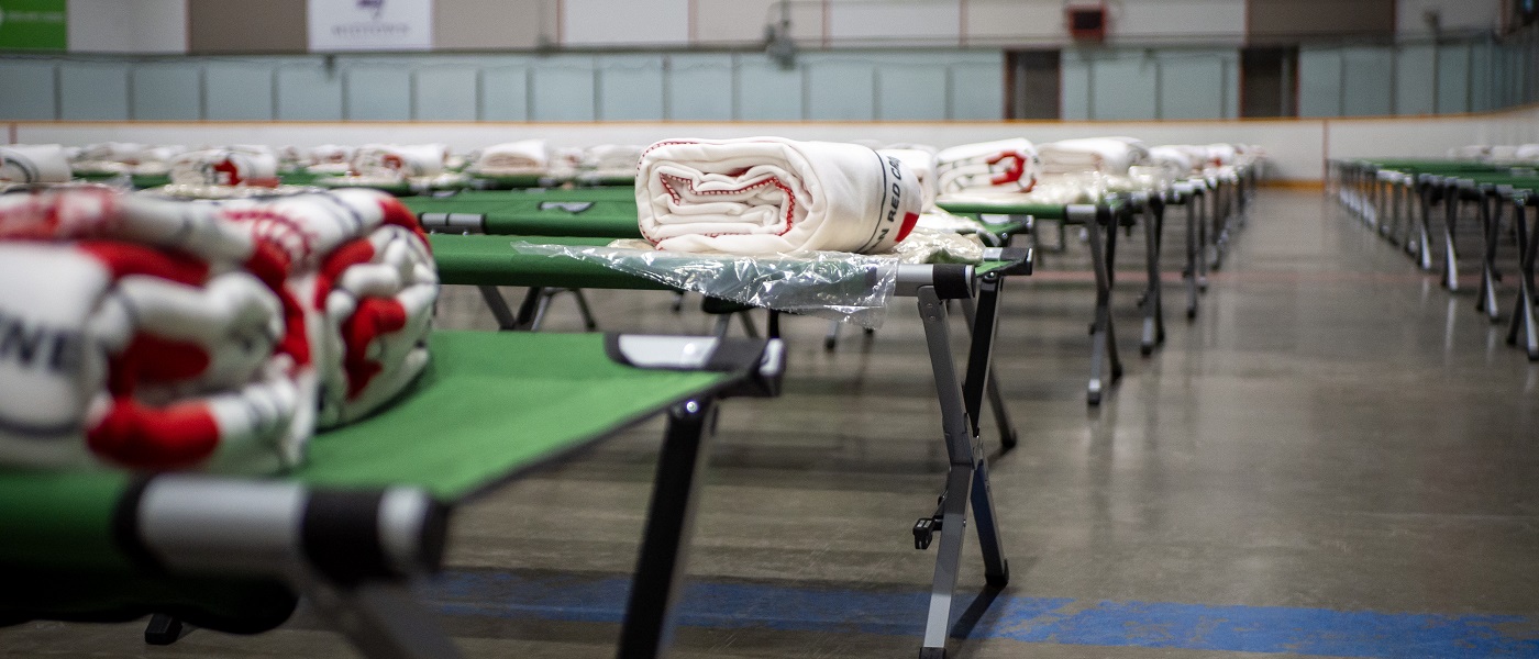 Rows of green cots with folded Red Cross blankets on them sit on the floor of a hockey arena. 