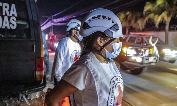 Venezuelan Red Cross personnel wearing helmets and masks stand near a vehicle at night during earthquake response efforts.