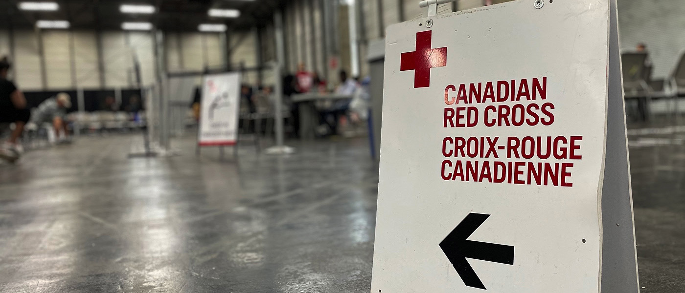 Une affiche dirige les gens vers les services de la Croix-Rouge canadienne offerts dans une grande salle, des gens patientent en arrière-plan.