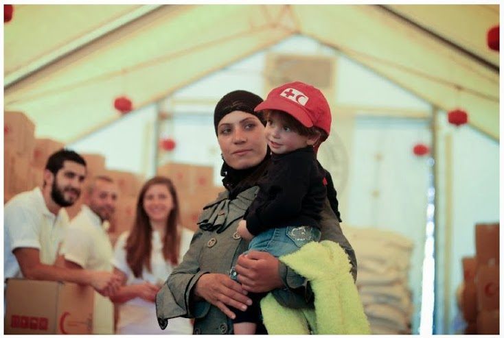 A woman holding a child, wearing a red hat with the IFRC logo.