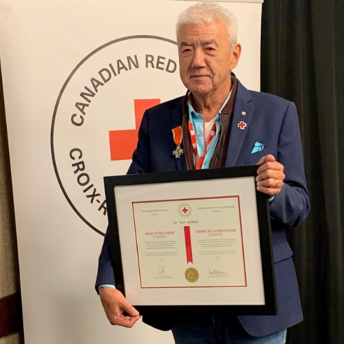 A man stands in front of a Canadian Red Cross Banner, holding an Order of the Red Cross award.