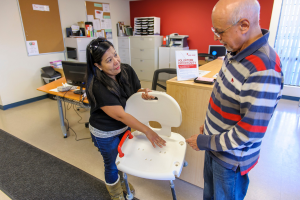 A person holds a shower chair, in discussion with another person.