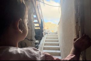 A young girl at the bottom of a staircase looks up and outside. There is a damaged wall on her left.