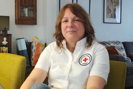Louise Bélanger wearing a white collared shirt with the Red Cross emblem, sitting on a chair in her home.