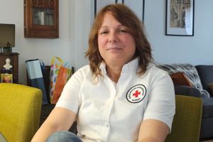 Louise Bélanger wearing a white collared shirt with the Red Cross emblem, sitting on a chair in her home.