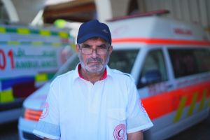 A headshot of Awni wearing a hat and glasses, standing in front of two ambulances.