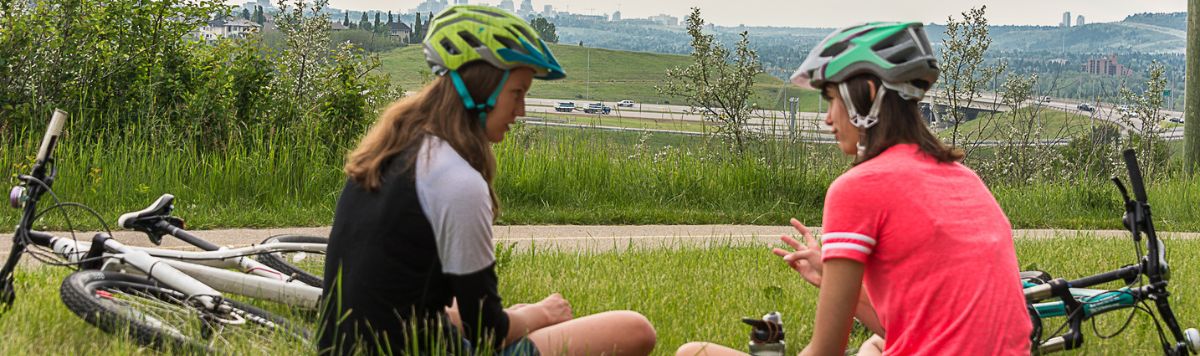 Deux jeunes personnes s’assoient sur la pelouse à côté de leurs bicyclettes. Elles parlent sans enlever leur casque, comme si elles prenaient une pause.