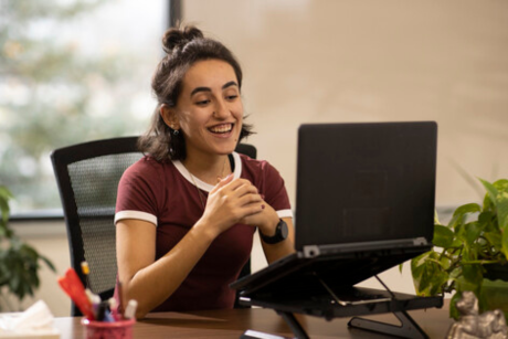 A person working remotely at home smiles at the camera on his desktop computer while clasping his hands together.