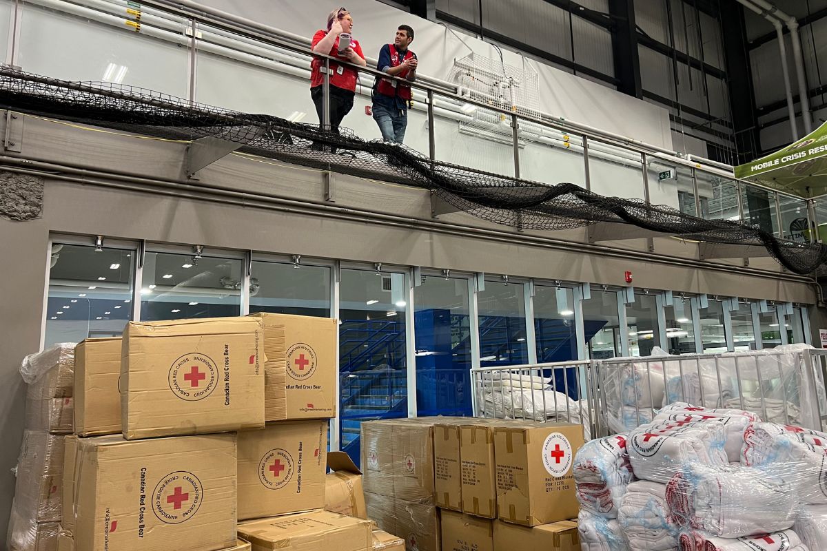Deux membres du personnel de la Croix-Rouge canadienne se tiennent sur une plateforme d’observation surplombant le matériel d’urgence et les couvertures disposées sur le sol du stade en contrebas.