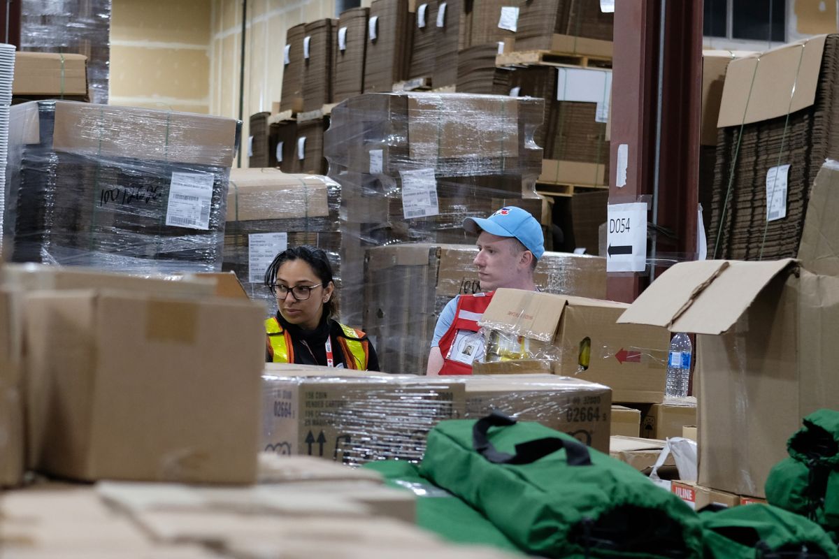 Dans un entrepôt, Fatima et Joshua sont entourés de boîtes de carton remplies de matériel.