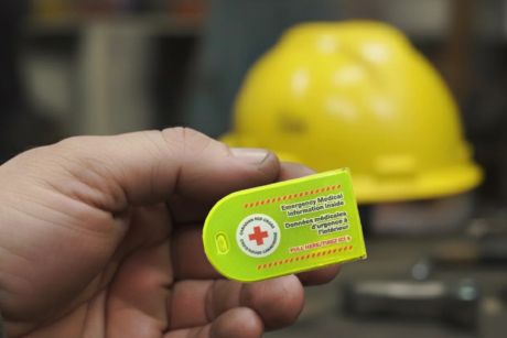 Une main tient une pochette de données médicales d'urgence de la Croix-Rouge. En arrière-plan, un casque de protection jaune est posé sur un établi.