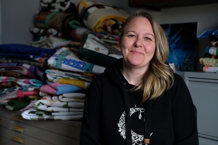 Photograph of a person sitting indoors, in front of a stack of quilts.