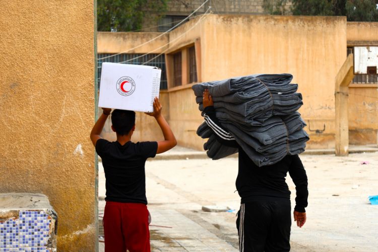 Two people carry supplies from the Syrian Arab Red Crescent through a village.