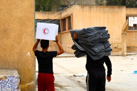 Two people carry supplies from the Syrian Arab Red Crescent through a village.