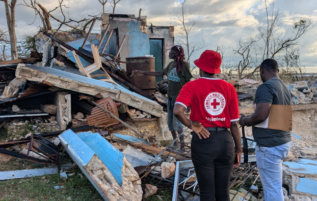 A red cross volunteer standing in front of a building destroyed by Hurricane Melissa