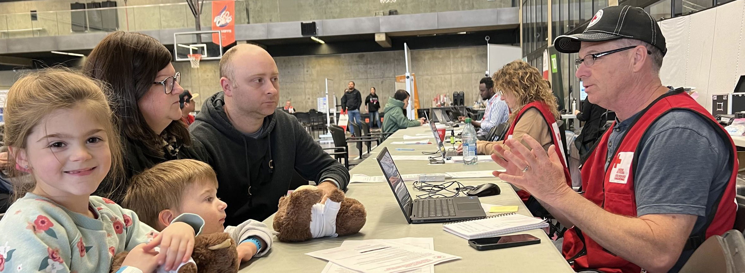 A young family of four sit across a table from a Red Cross volunteer, having a conversation. Their young daughter smiles at the camera as she hugs a teddy bear.