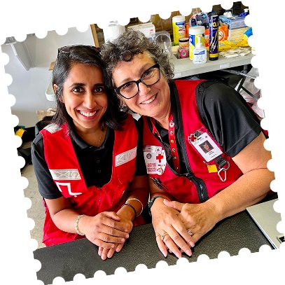 Two female Red Cross volunteers wearing red vests, sitting side by side with big smiles on their faces.