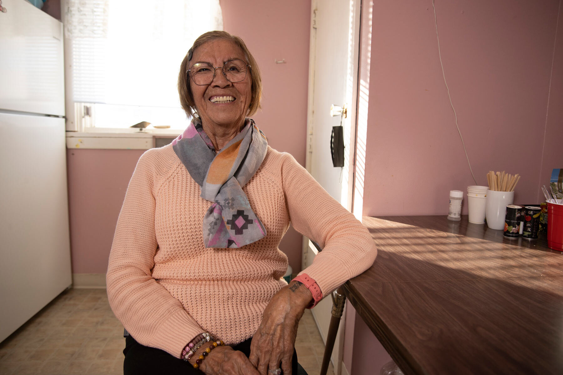 An elderly woman wearing a pink sweater and silk scarf sits in her sun-lit kitchen, smiling.