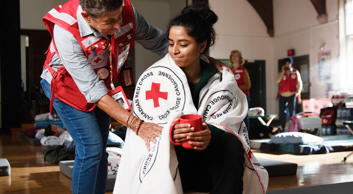 A woman holds a red mug as a Red Cross volunteer wraps her shoulders in a blanket. The interior is an indoor emergency shelter.