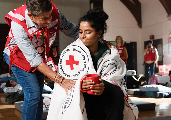 A woman holds a red mug as a Red Cross volunteer wraps her shoulders in a blanket. The interior is an indoor emergency shelter.