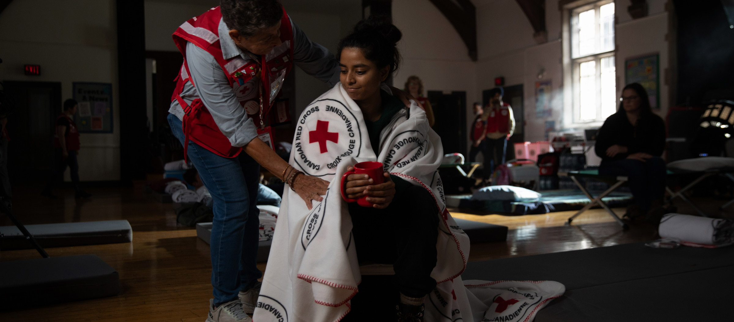 A woman holds a red mug as a Red Cross volunteer wraps her shoulders in a blanket. The interior is an indoor emergency shelter. 