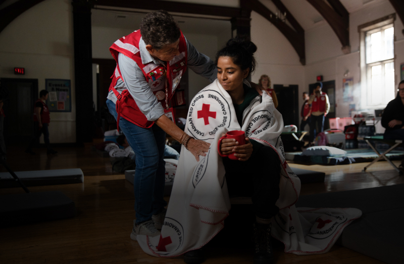 A woman holds a red mug as a Red Cross volunteer wraps her shoulders in a blanket. The interior is an indoor emergency shelter. 