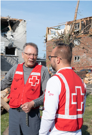 In front of a ruined building, two men in red cross vests engage in conversation after tornadoes.