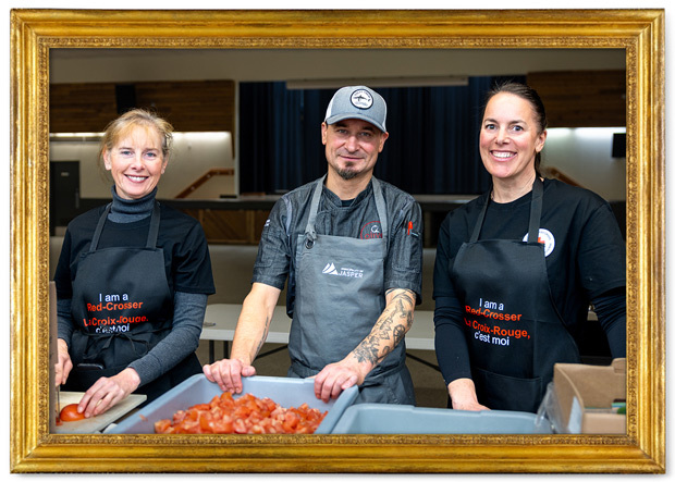Three smiling kitchen volunteers behind a table with a bin of chopped tomatoes