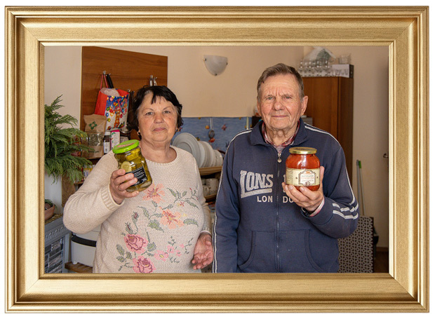 A man and woman pose for a photo, each holding a jar of preserved food (pickles and tomato sauce). 
