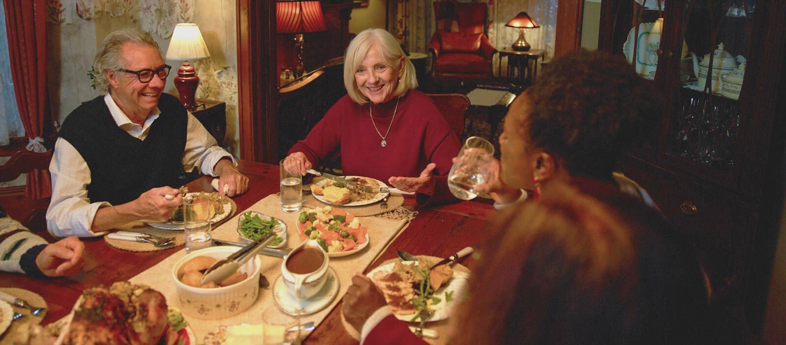 Two smiling adults, a man in glasses and a woman in a red sweater, sitting around a dining table, sharing a holiday meal. 