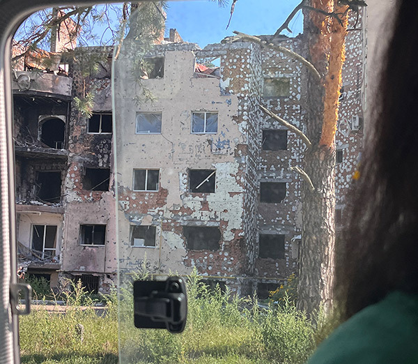 A person looks out a vehicle window at destroyed buildings.