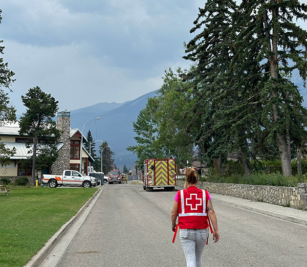 A Red Cross worker walks down a street with a firetruck in the distance.