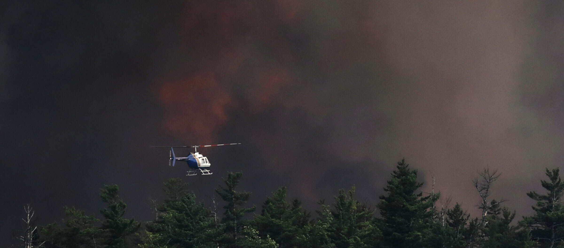 A helicopter hovers over a forested area overtaken by clouds, smoke and flames in Nova Scotia.