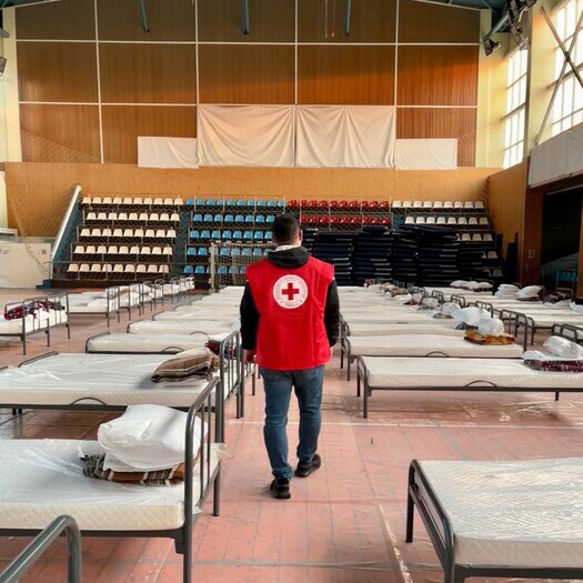 Volunteer in a red Canadian Red Cross vest walking down a row of beds, engaged in assistance and care activities. 
