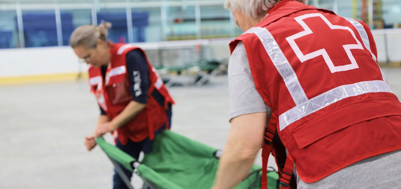 Two Red Cross volunteers in red vests stand on an indoor ice rink, working together to open and set up a green cot.