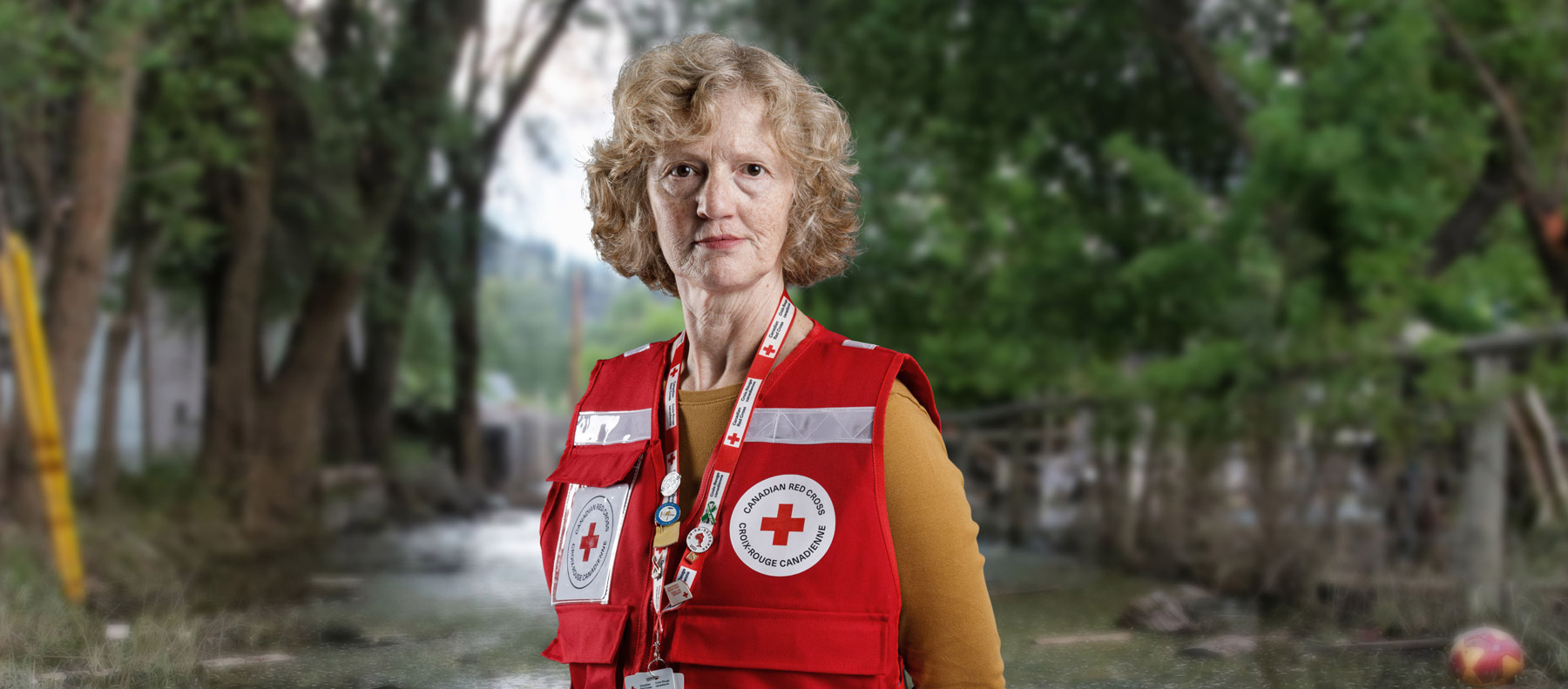 Une bénévole plus âgée de la Croix-Rouge porte un gilet rouge décoré de broches de la Croix-Rouge. Elle se tient devant un parc inondé et son visage est préoccupé. 