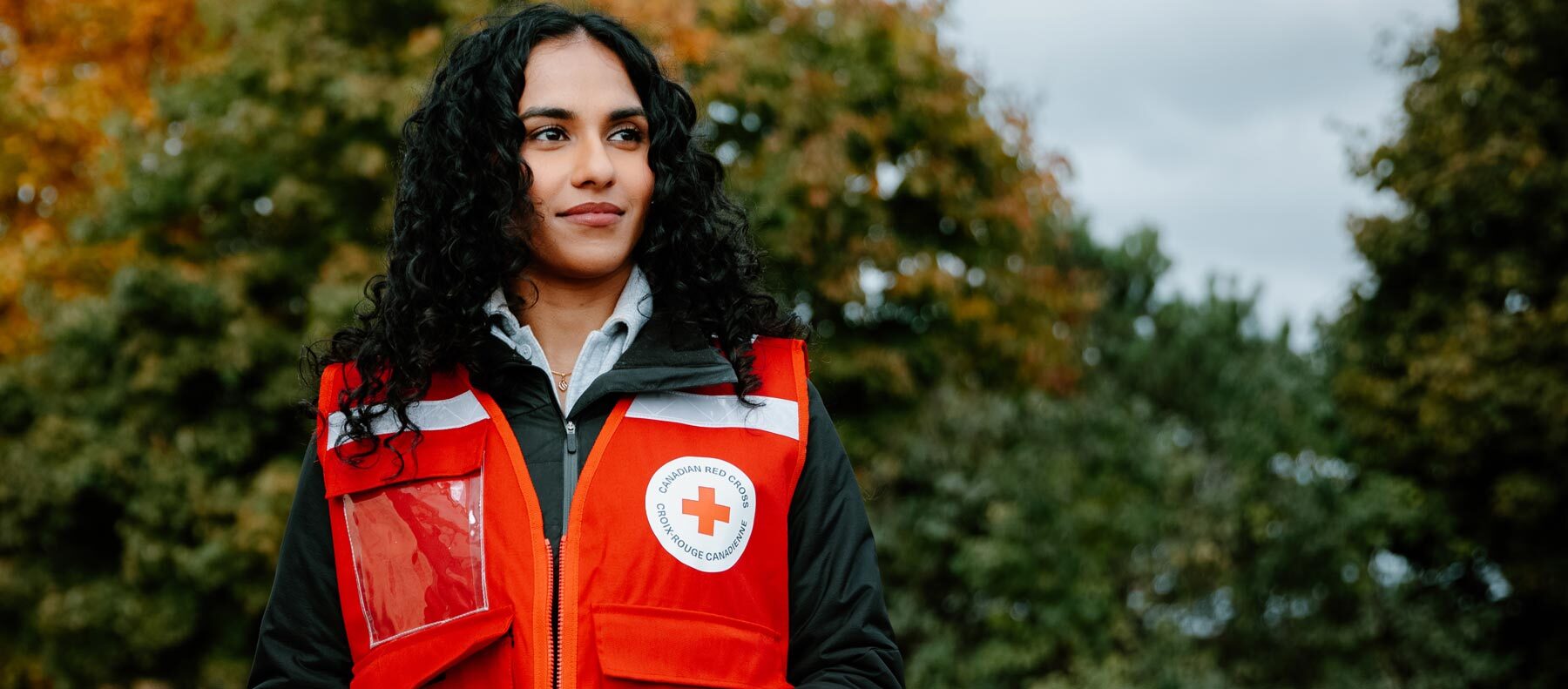  A woman in a Canadian Red Cross vest stand in front of backdrop of green trees.