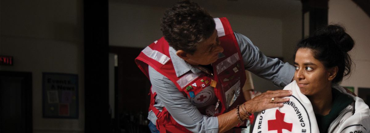 Red Cross personnel comforting a woman who has a white Red Cross blanket wrapped around her.