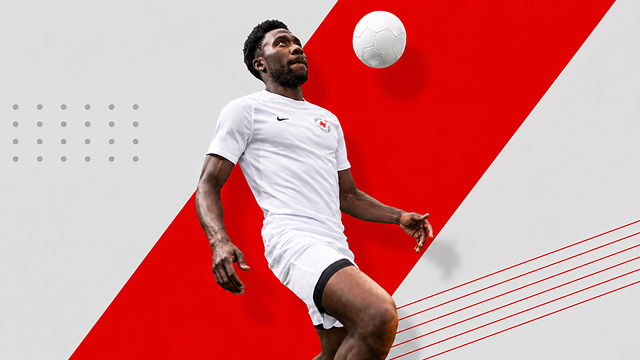 Alphonso Davies, concentré, donne un coup de pied dans un ballon de soccer en l'air, vêtu d'un t-shirt blanc de la Croix-Rouge. 