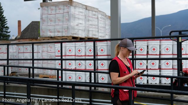 Canadian Red Cross personnel using a cellphone, standing in front of humanitarian supplies.