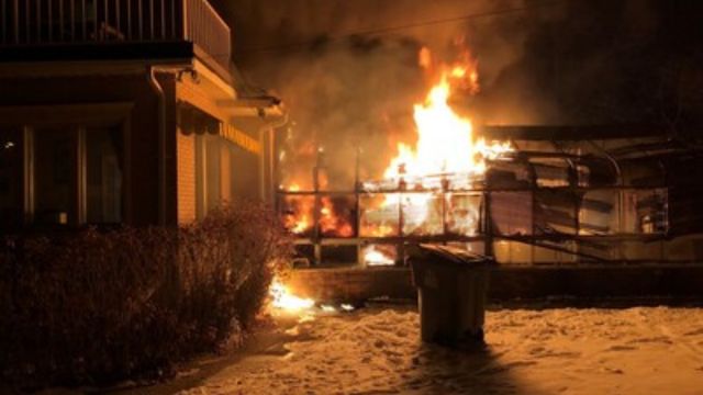 A shed behind a house on fire at night, with snow on the ground and firelight illuminating the space