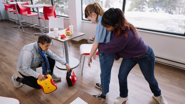First aid training in a cafeteria, with one participant bringing an AED while another demonstrates abdominal thrusts on a standing participant.