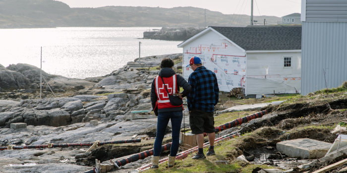 Deux personnes, debout, discutent près d’un plan d’eau. Une personne porte un dossard de la Croix-Rouge canadienne. En arrière-plan, un édifice est en construction. 