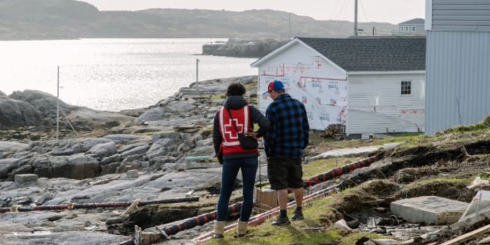 Two people stand near the water talking. One person is wearing a Canadian Red Cross vest. There is a building under construction in the background. 