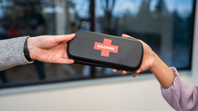 The hands of a person handing a Naloxone kit to another person. 