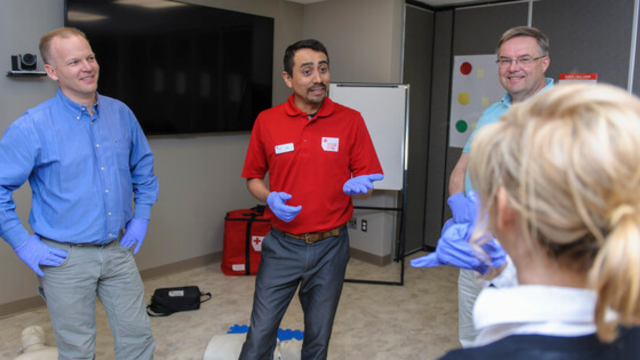 Person wearing a Red Cross Instructor shirt standing in a classroom speaking to two learners.